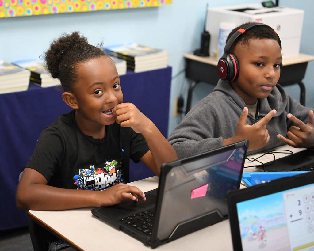 Two children at a desk with laptops. One has headphones and is making gestures, the other is smiling.