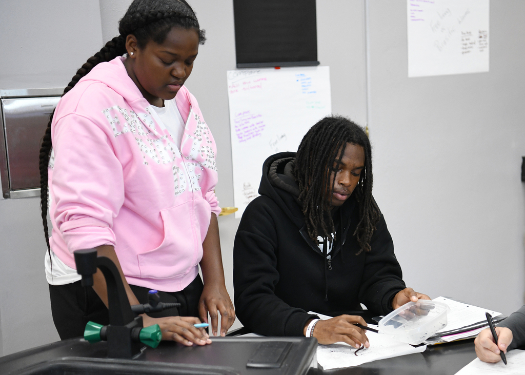 Two people seated at a table with papers and pens. A woman in a pink hoodie and a man in a black hoodie.