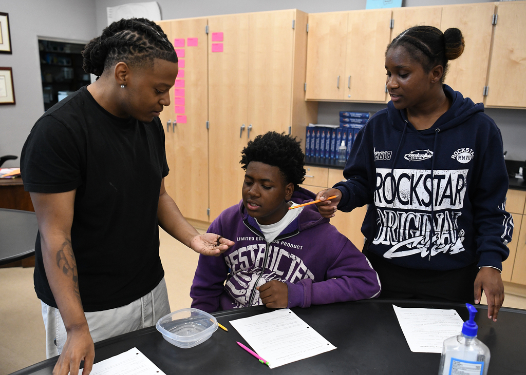 Three people are in a classroom with cabinets and papers on a table. One person has a toothbrush in another person's mouth.