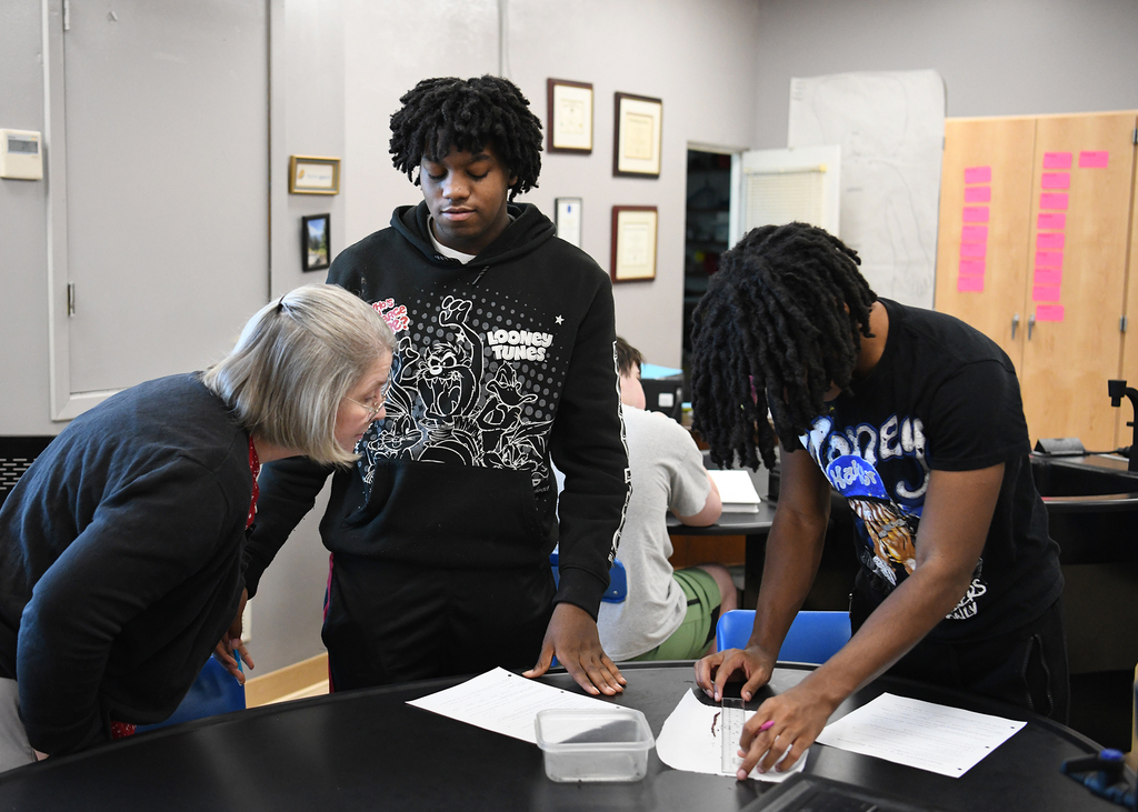 Three people in a room with gray walls, examining papers on a black table. One person wears a hoodie.