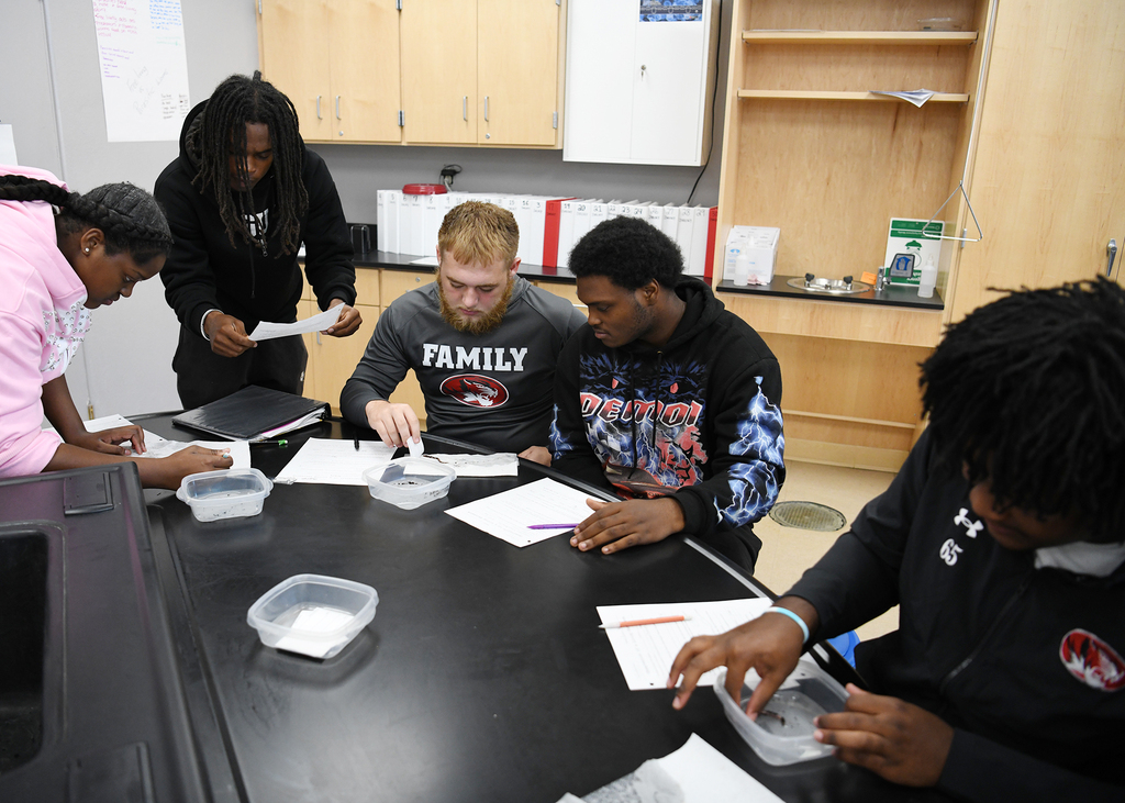 Five students with various hairstyles are at a table, looking at papers and plastic containers.