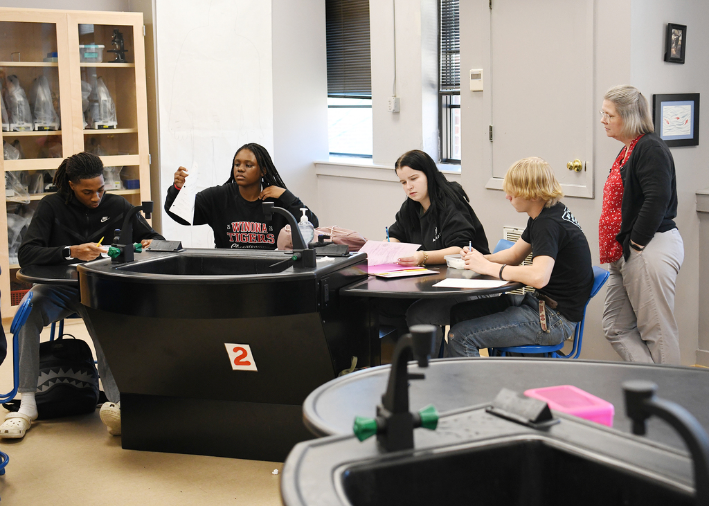 Five students sit at a lab table, one gesturing, others writing. The teacher stands nearby, and the room has lab equipment and a glass cabinet.