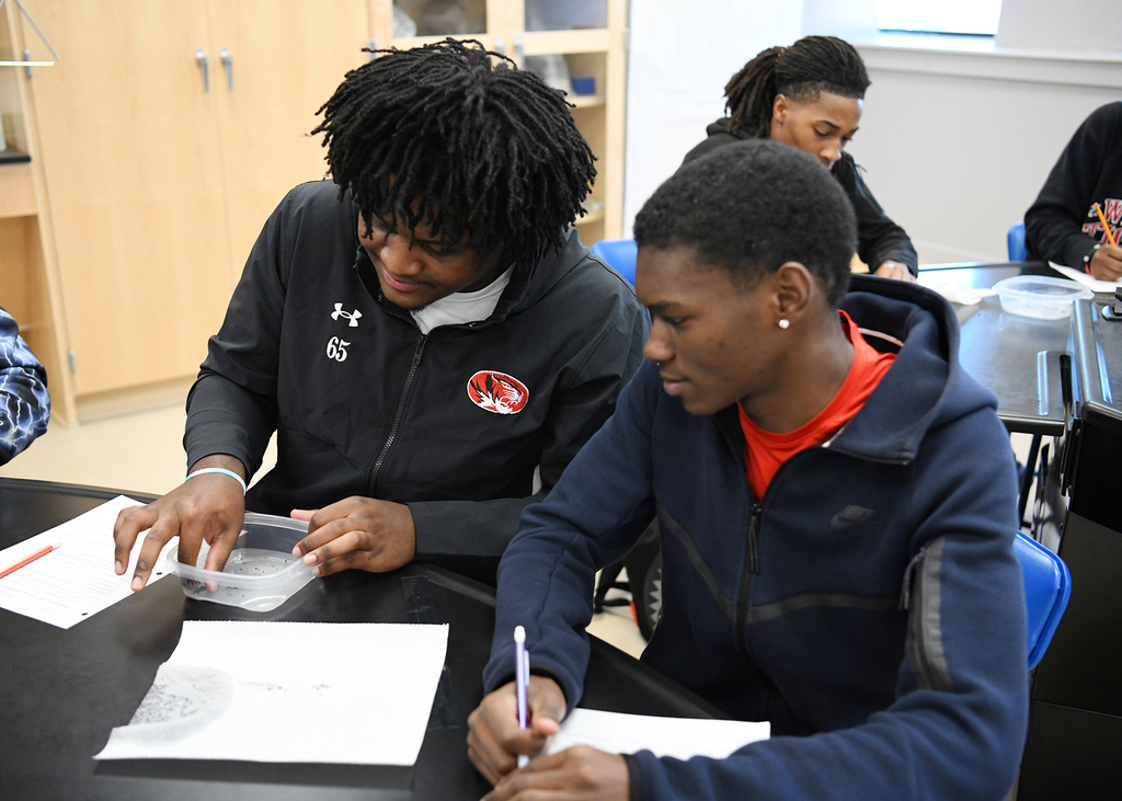 Two students work on a science project at a table, using paper and a pen. One student has a plastic container.