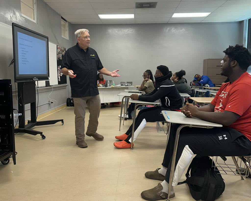 A man stands in front of a classroom with a screen, gesturing while explaining. Students sit at desks.