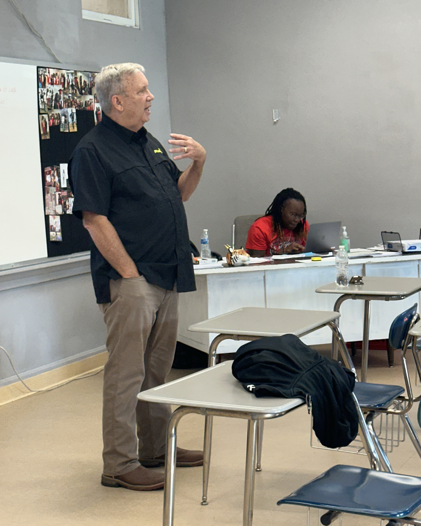 A man in a black shirt stands in front of a classroom, gesturing with his hands. A student sits at a desk in front of him.