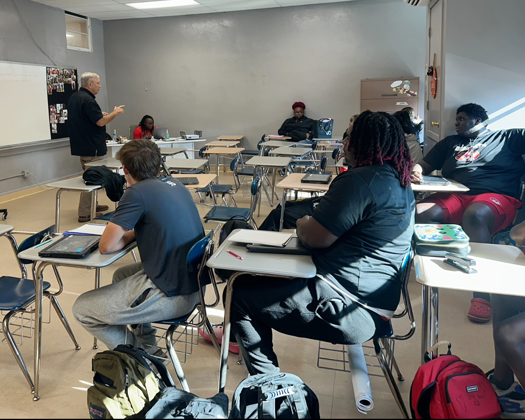 A classroom with students and a teacher. Desks and chairs are in rows. A white board is on the wall.