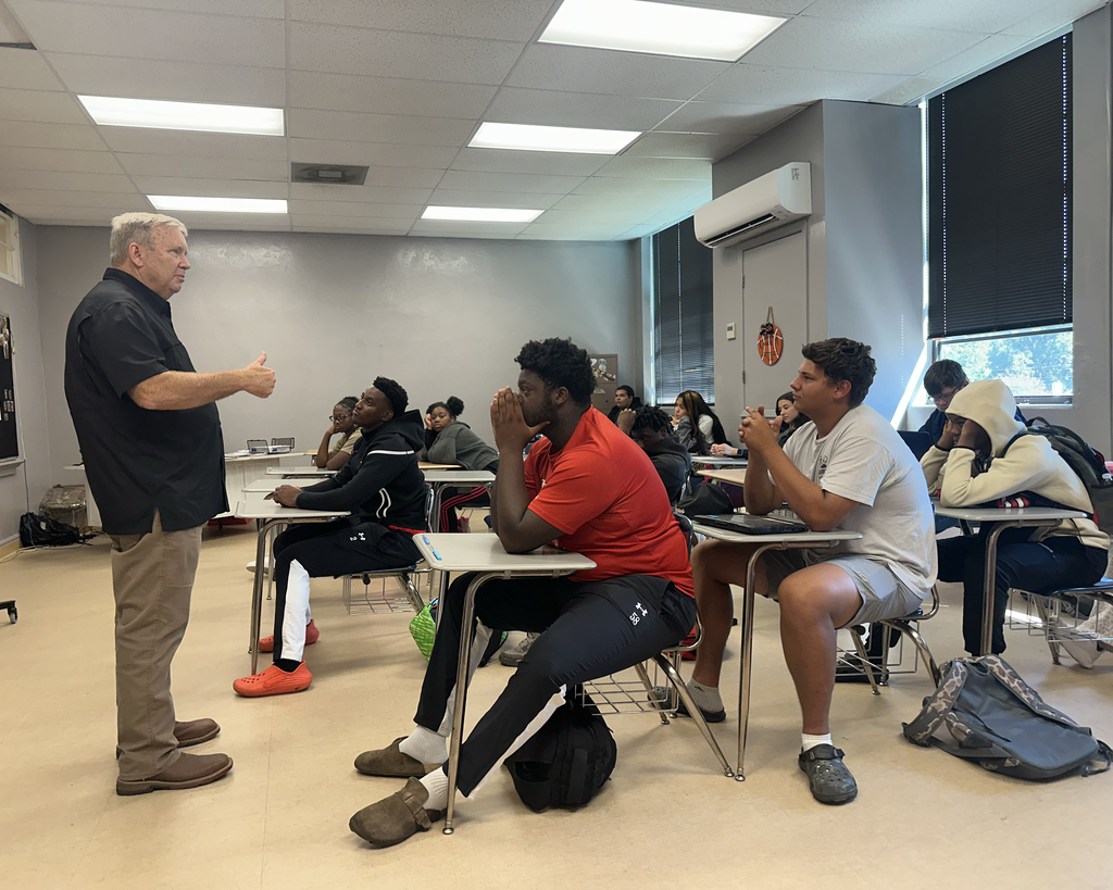 An instructor stands in a classroom, addressing a group of students seated at desks, with backpacks and notebooks visible.