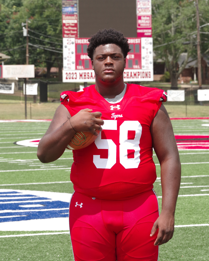 A football player in red uniform holds a football, standing on a field with a "2023 MHSAA CHAMPIONS" sign.