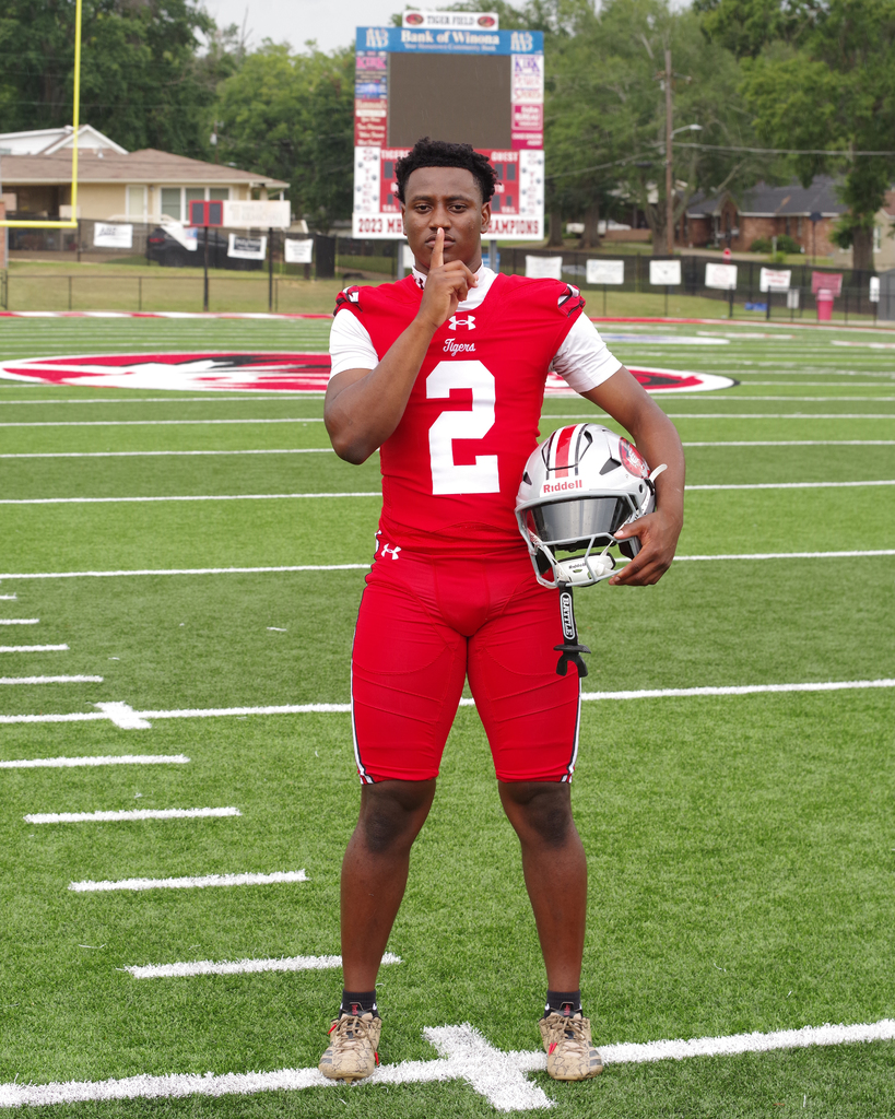 An athlete in a red uniform stands on a football field holding a helmet. Behind them, a scoreboard displays information.