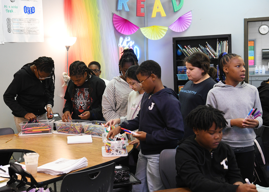 Children gathered around a table with craft supplies, standing in a room with rainbow decorations and a bookshelf.