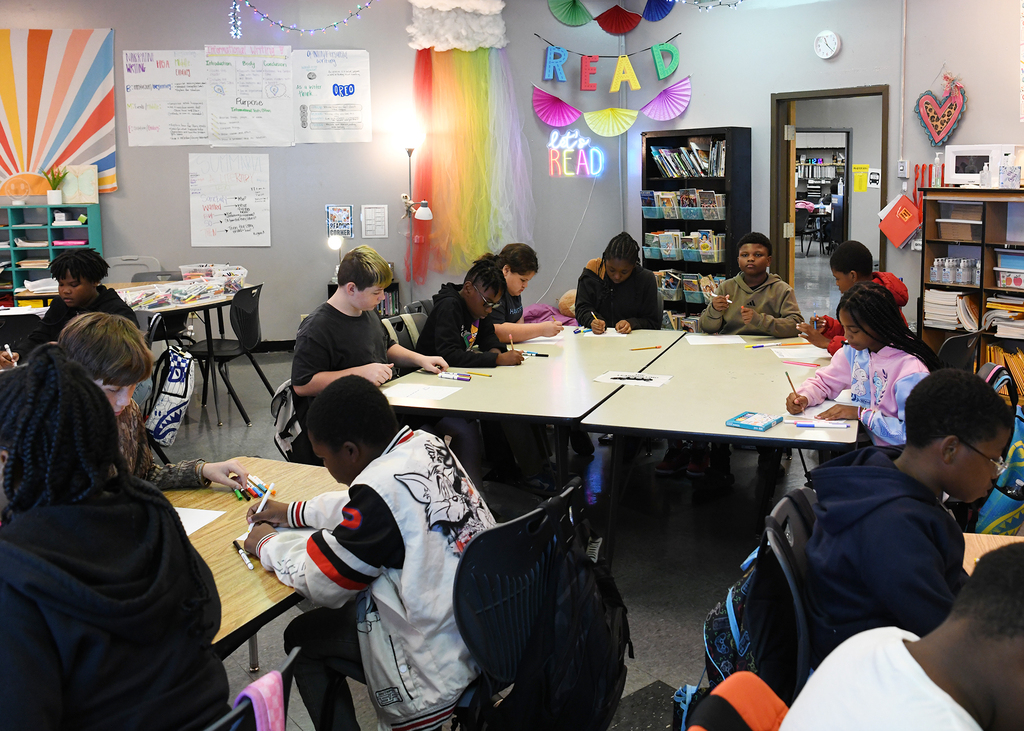 Classroom with tables; students sit around tables, engaged in writing activities; walls have decorations and boards.