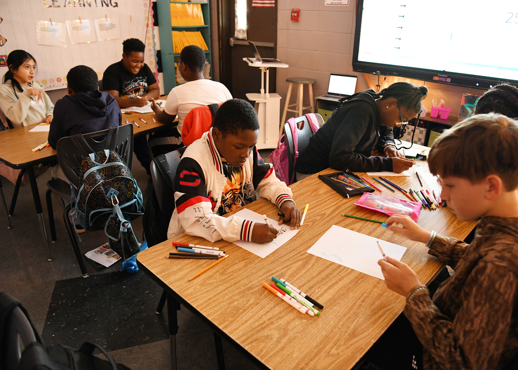 Students sit at desks in a classroom, facing a large screen. Some hold pens and papers.