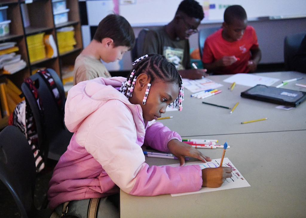 Children in a classroom; one girl in a pink jacket is writing, others are looking at papers.