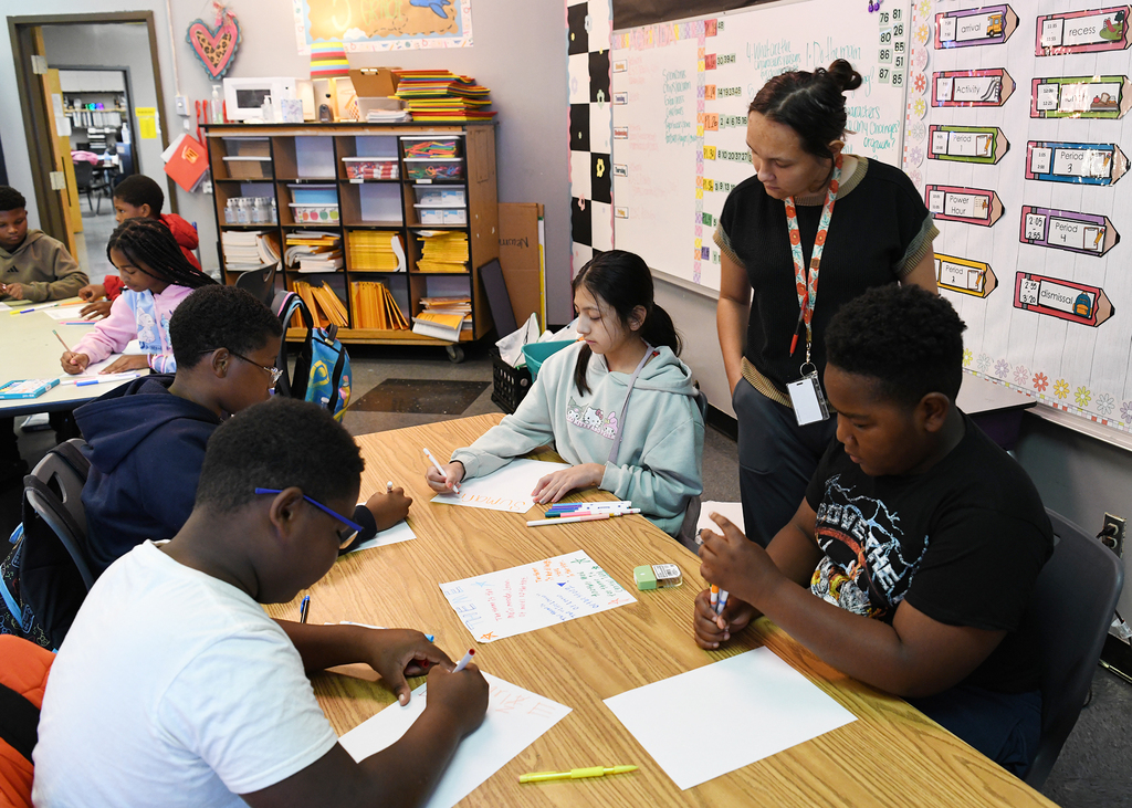 Students in a classroom are working on papers. One student is sitting with papers, and a teacher is standing behind them.