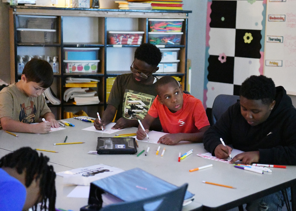 Five children sit around a table with writing tools and papers. They are focused on their activities.
