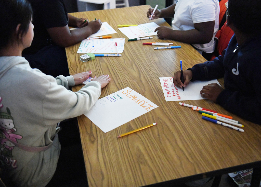 Four people around a table with papers, pencils, and markers. One person wears a white sweatshirt, another wears a blue sweatshirt.