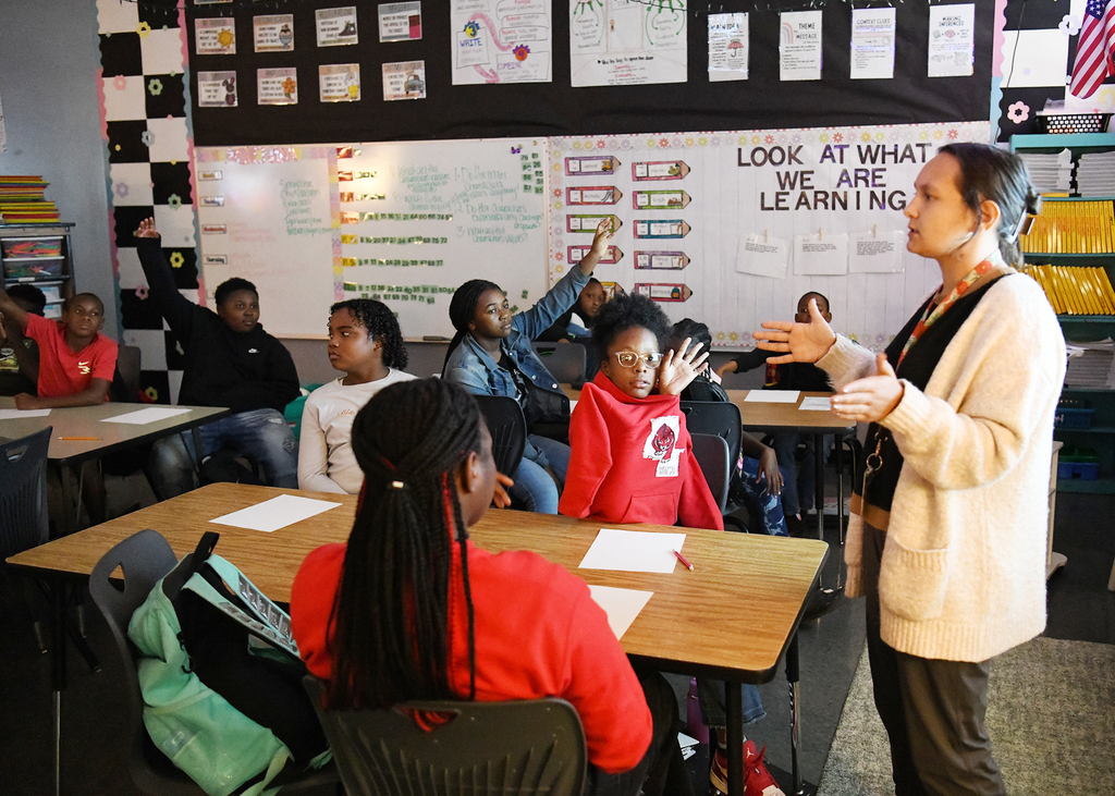 A teacher stands in front of a classroom with a whiteboard, teaching a group of students.