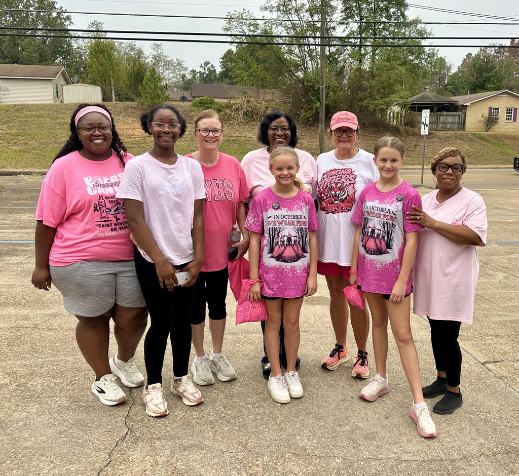 A group of people stand in a parking lot, wearing pink shirts. Trees and houses are in the background.