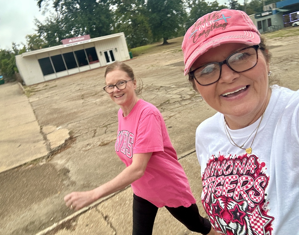 Two women in pink shirts and caps walk in a parking lot, with a white building in the background.