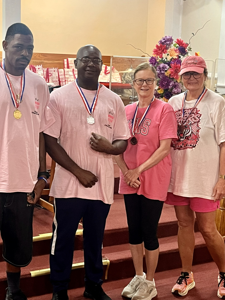 Four people in pink shirts and shorts stand on stairs with medals. Two men wear black pants, one wears shorts. Two women wear glasses and caps.