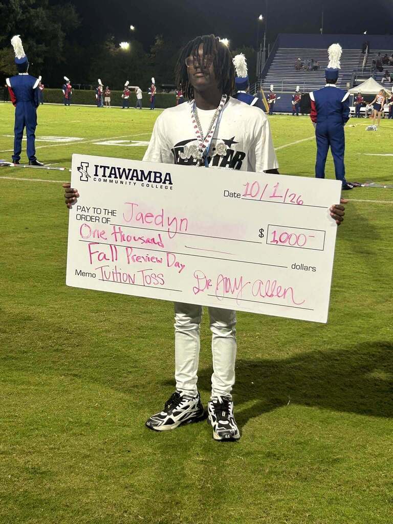 Person stands on a football field holding a large check for $1,000. Behind them, band members are in blue uniforms.