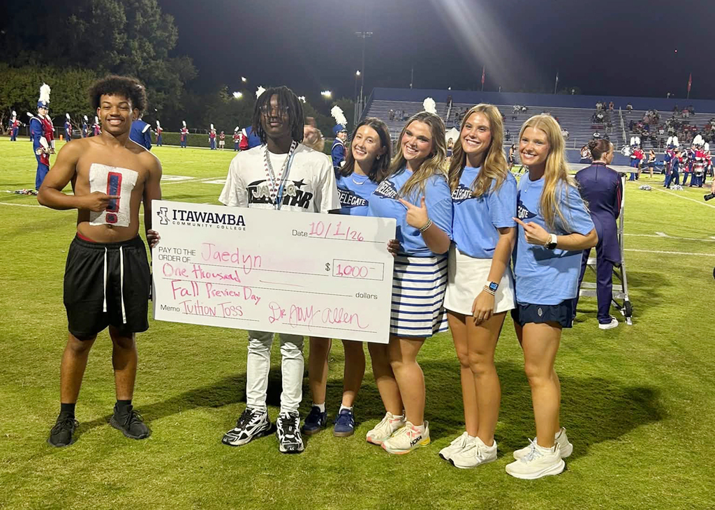 Five people holding a large check on a sports field. Three women and two men, one wearing a jersey.