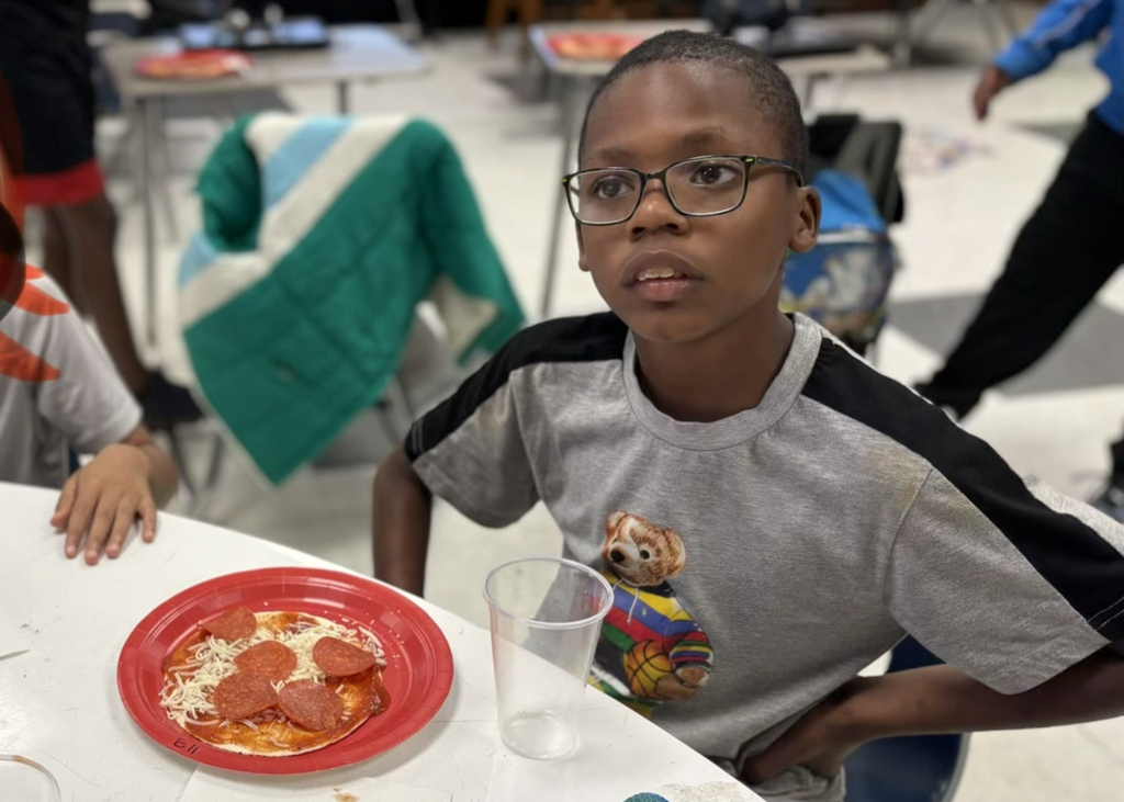 A boy with glasses eats pizza at a cafeteria table. Another child sits behind him.