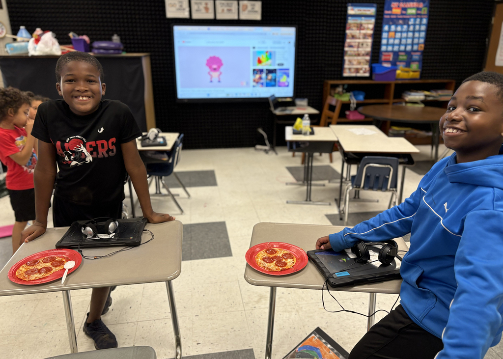 Two kids in a classroom sit at desks, facing each other. They both have a plate of pizza.