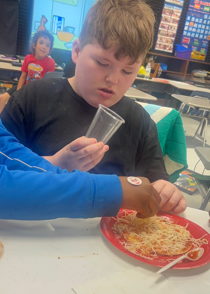 Boy eating pizza on a red plate. Another person holds a plastic cup. Brightly lit classroom background.