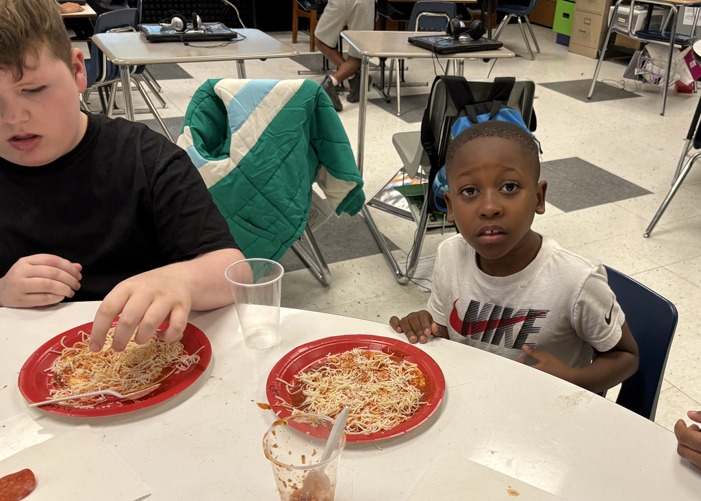 Two kids eating pizza at a table in a classroom. One wears a black shirt, the other a white shirt.