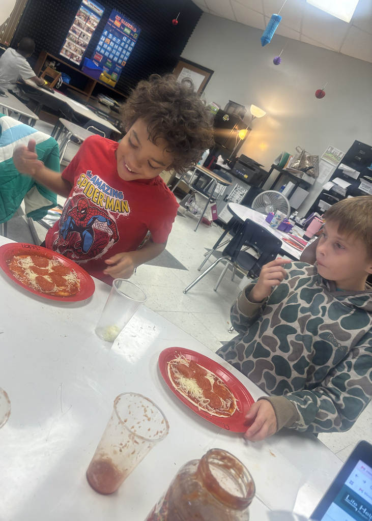 Two children in a classroom eating pizza on red plates; one boy wearing a red shirt.