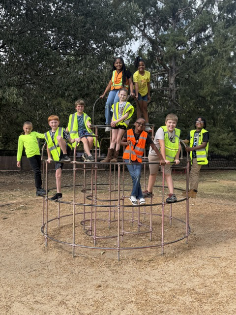 A group of kids in bright vests, posing on a climbing frame in a park with trees in the background.