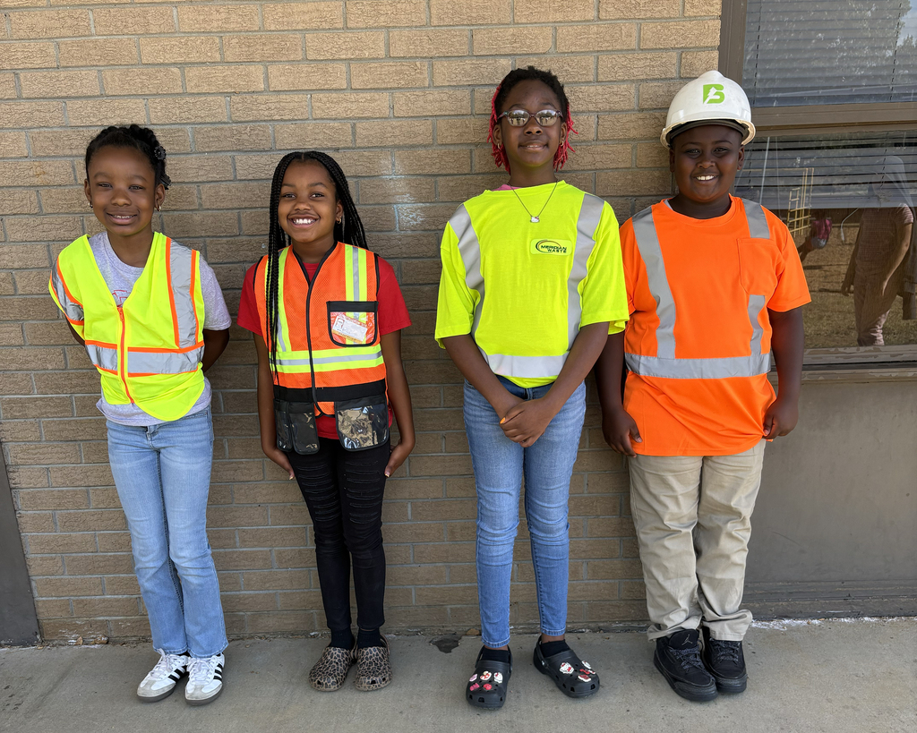 Four kids in reflective vests stand against a brick wall, likely for a photo. They wear different colors of vests and sneakers.