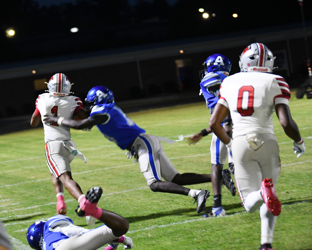 Football players wearing helmets and sneakers, one tackling another on the field at night.