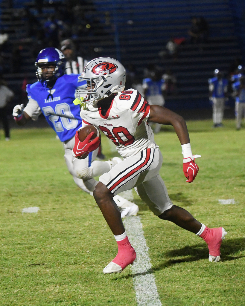 A football player runs with a ball, wearing a gray helmet and white jersey. Another player in blue chases behind.