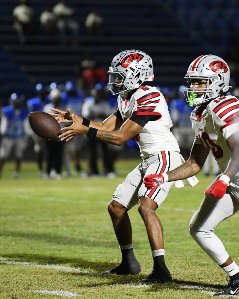 Two players in white uniforms and helmets on a field, one holding a football while the other looks on.