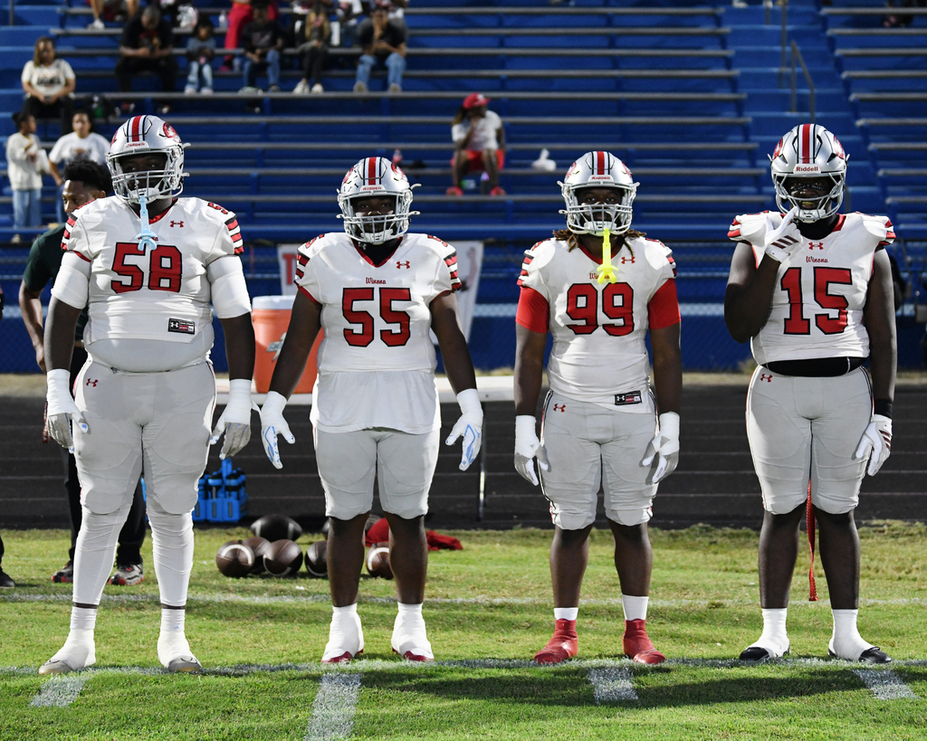 Four football players in white jerseys and helmets, numbers 58, 55, 99, and 15, standing on a field with bleachers in background.