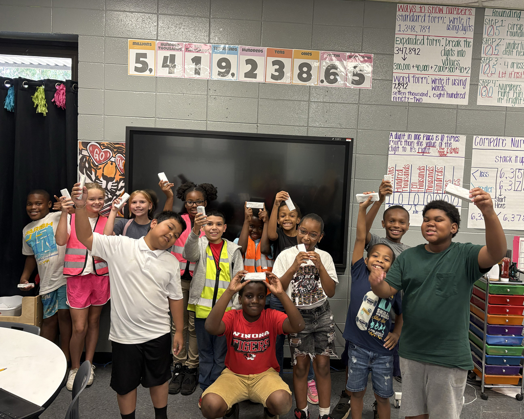 A group of children in a classroom holding Wii remotes, standing and sitting in front of a blackboard.