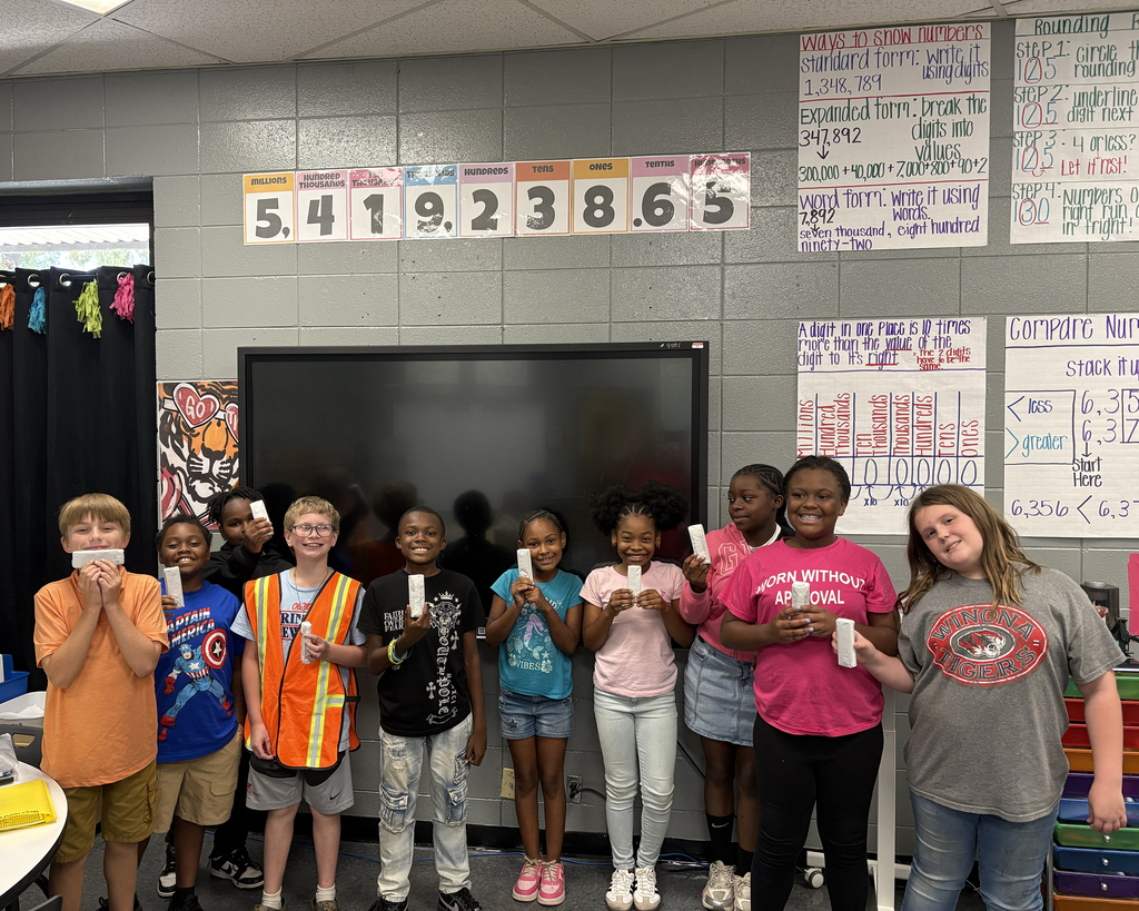 Nine children and two adults stand in a classroom. The children hold up a white object.
