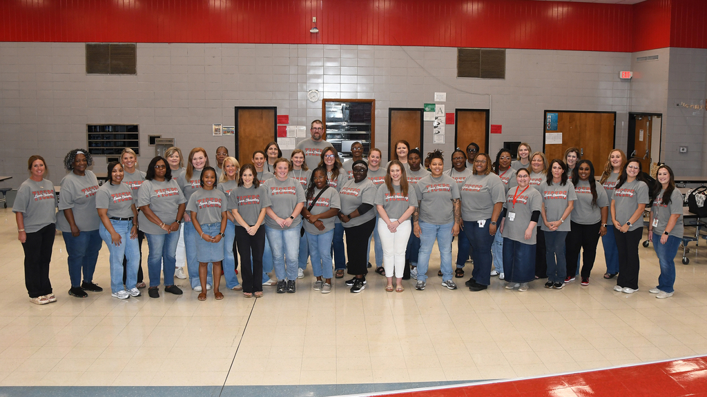 Group of people in matching gray shirts and jeans stand in rows, with a red-and-white wall and doors behind.
