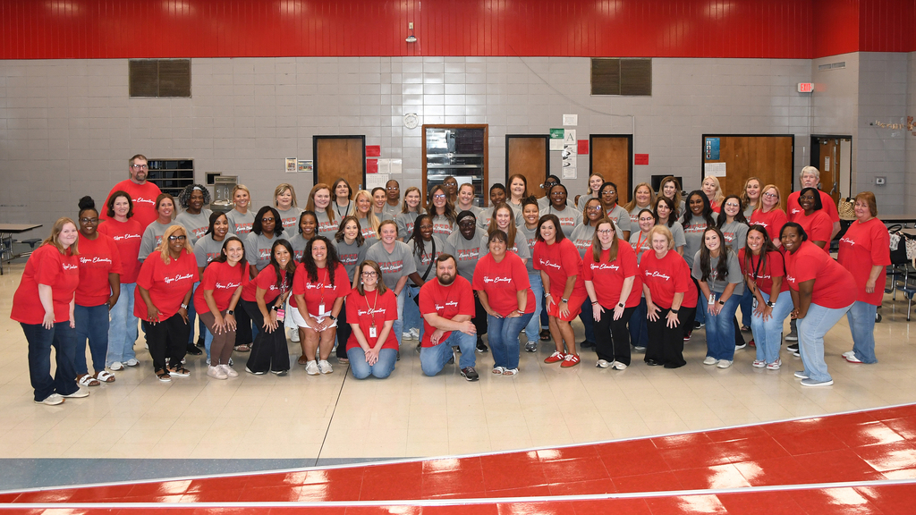 Group of people wearing red shirts and blue jeans, posing for a photo in a gymnasium.