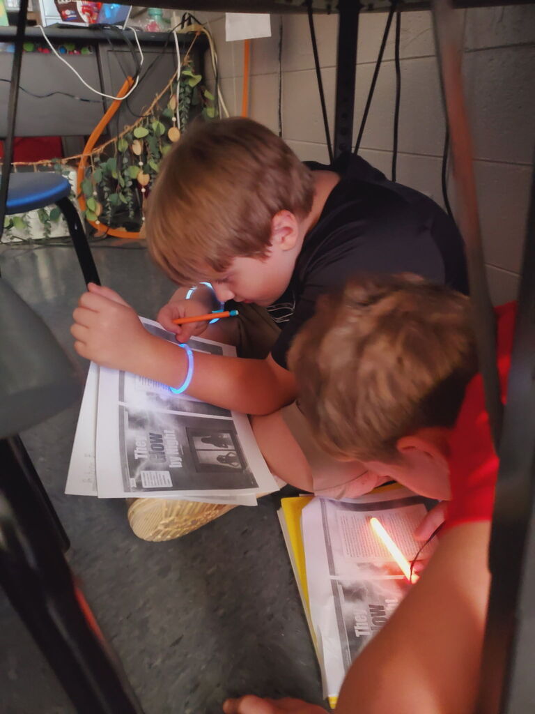 Two boys are seated on the floor, focusing on papers, one holding a pen and the other a flashlight.