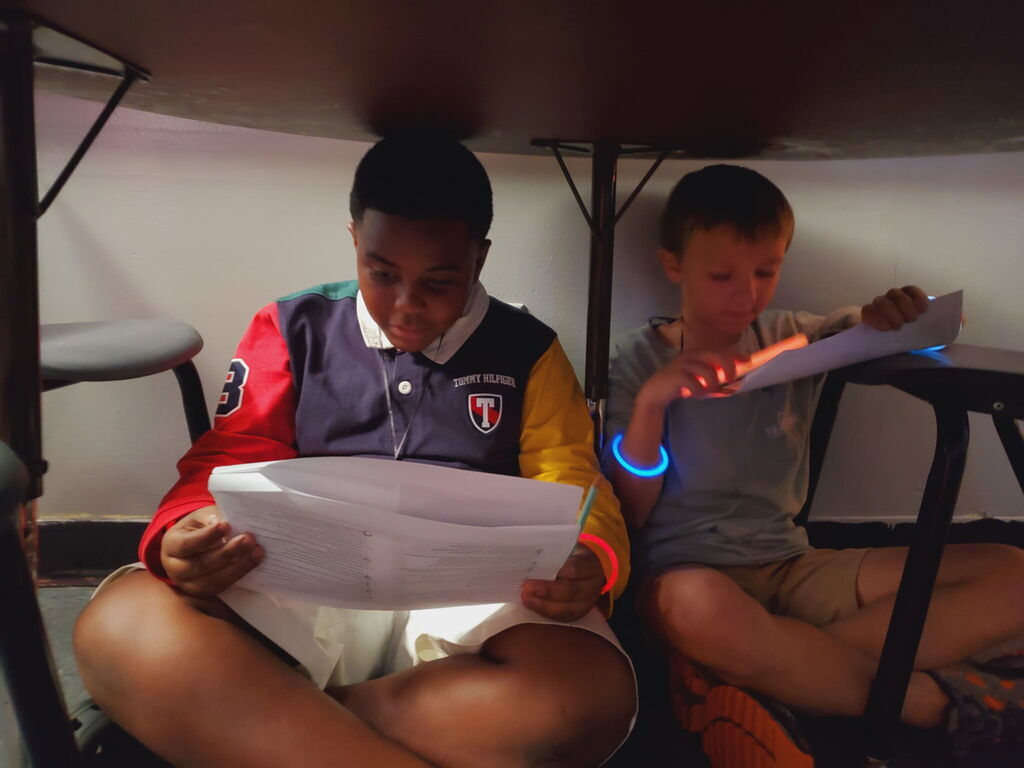 Two young people sit under a table. One reads a paper, and the other reads a digital tablet.