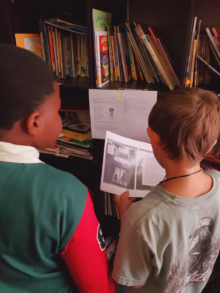 Two boys face each other while looking at a book in a library filled with books.