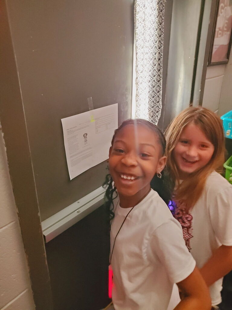 Two girls with short hair stand close, smiling at camera. One wears a necklace, other wears a ribbon. Behind them is a wall with a bulletin board.