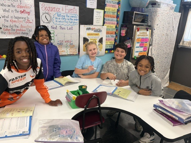 Five young students, wearing jackets, sit around a round table with papers and books, in a classroom.