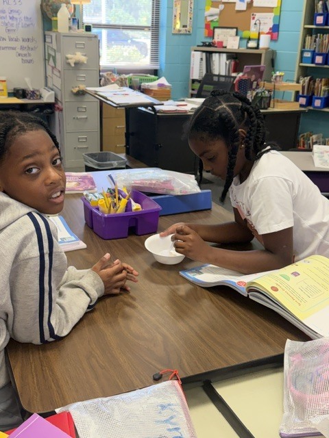 Two students seated at a table, one looking at the other, who holds a bowl and a book.