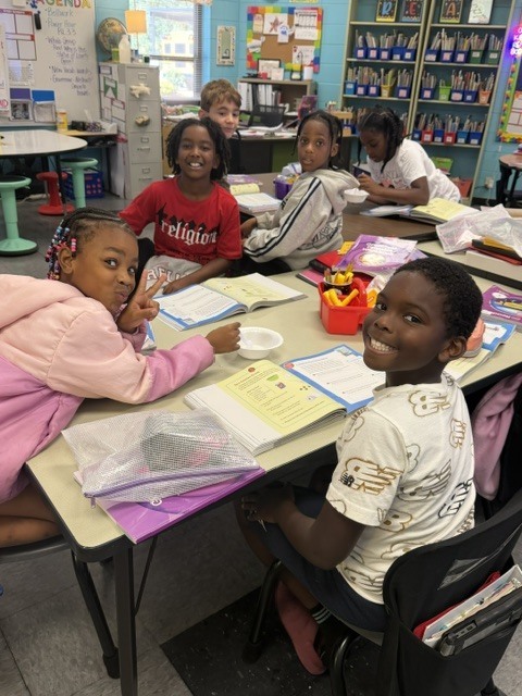 Children are seated at a table with books and supplies in a classroom. Some wear jackets.
