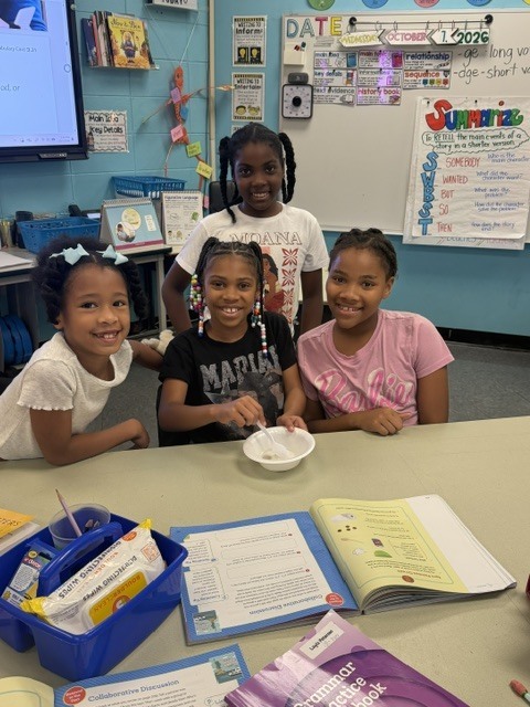 Four girls in a classroom are seated around a table with a bowl and various books.