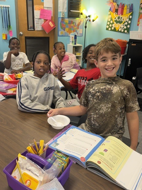A classroom with children sitting at a table. A boy holds a book and smiles. Other children look on.